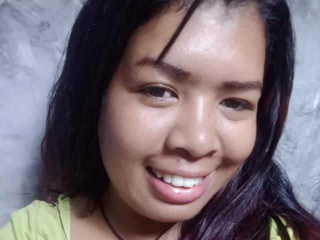 Close-up photo of MariahSquirting69 with medium brown skin and straight black hair with a slight red tint. She has dark brown eyes, thick eyebrows, and a small nose. MariahSquirting69 is smiling with visible white teeth. She wears a light green shirt with a dark necklace. The texture of her skin is smooth with a slight sheen on her forehead and nose. The image has a soft, slightly blurry background. The photo is centered on her face, focusing on her facial features and expression.