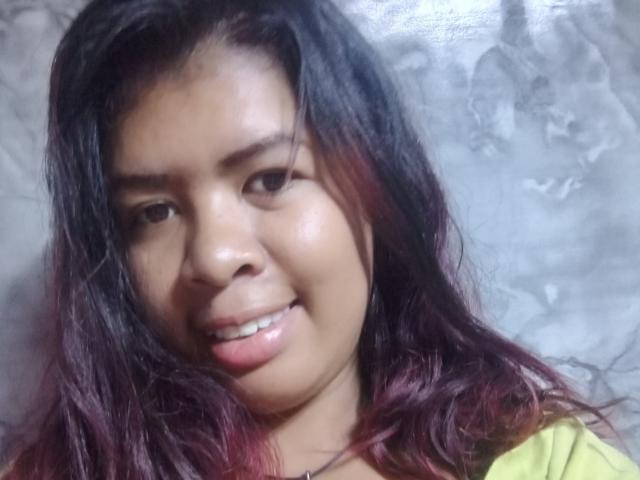 Close-up photo of MariahSquirting6. She has medium-dark skin, black wavy hair with red highlights, and a slight smile showing her teeth. Her eyebrows are thick and she has a small nose. She wears a yellow top with a dark necklace. The light is soft, highlighting her natural skin texture. The background is a textured, gray surface. Her hair is slightly messy, and her expression is relaxed. The image focuses on her face and upper shoulders.