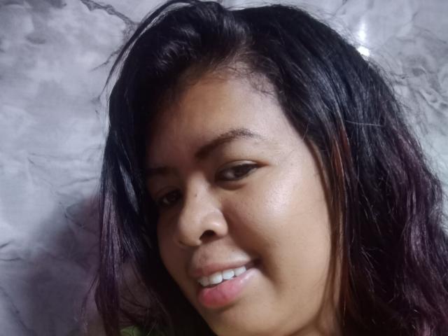 Close-up photo of MariahSquirting6. She has medium brown skin, black straight hair with purple highlights, and subtle black eyeliner. Her lips are slightly parted with a small smile. She has small, dark eyebrows and a slight shadow under her eyes. The background is a marble-patterned wall. The light is soft, highlighting her facial features. The image is clear and focused on her face.