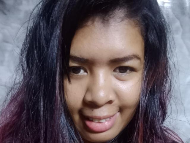 Close-up photo of MariahSquirting69. She has medium brown skin, dark brown eyes, and a small, slightly upturned smile. Her black hair with purple highlights is messy and falls around her face. She has a small nose and slightly parted lips, revealing a hint of teeth. The background is blurry and grey, keeping focus on her face. Her skin has a natural sheen, and her eyebrows are slightly raised. The lighting is soft, highlighting her facial features.