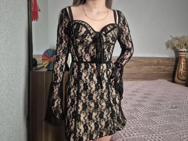 ZoieHurt wears a black lace dress with a deep V-neck, showing her small boobs, and black straps over her shoulders. Her hands rest on her hips.