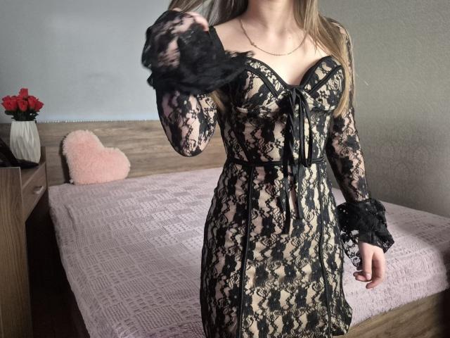 ZoieHurt wearing a black and beige lace dress, showing her tits, with black lace cuffs, standing in a bedroom with a pink bed and red roses on a nightstand.