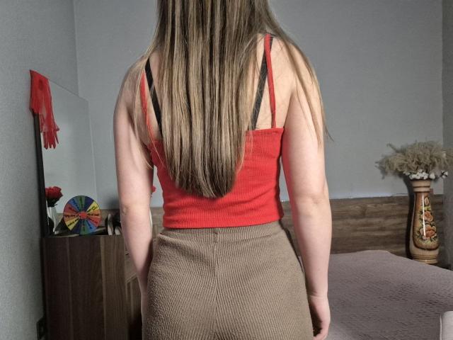 ZoieHurt met lang bruin haar, een rood tanktop en een bruke rok, waarbij zwarte bh-bandjes zichtbaar zijn, staat met haar rug naar de camera, in een kamer met houten meubels..