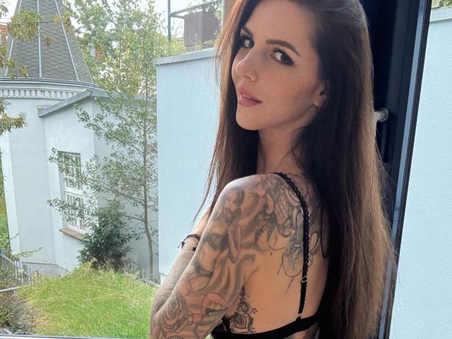 Sofialove6-hot with long brown hair, looking over shoulder, black lace bra, tattooed arm, standing by window, showing off her tits, with a slight smile, outside in daytime.