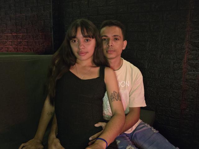 MadissonAndThomas with long brown hair in a black dress, sitting next to Thomas with short brown hair in a white tee, arm tattoos, on a dark brick wall.