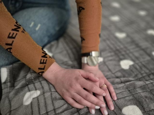 LoriEla's hands with white nails and a gold ring are touching a grey blanket with white heart patterns, wearing a brown sleeve with "ALEX" and a silver watch.