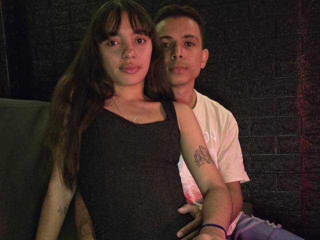 MadissonAndThomas with long brown hair, black sleeveless top, and small tits, sits next to Thomas with short brown hair, white tee, and tattooed arm, in dim lighting.
