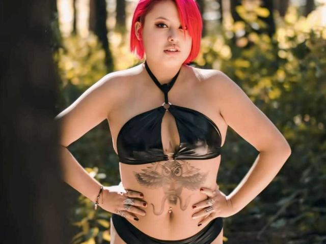 A photograph of RubiBabee with bright red, short hair standing in a forest. She wears a black, shiny, halter-style bikini top with a central cut-out, revealing a tattoo of a detailed, symmetrical eye with wings on her lower abdomen. RubiBabee's hands rest on her hips, showcasing black-painted nails and several rings. The sunlight filters through the trees, casting a warm, natural glow on her light skin. The forest background is blurred, emphasizing RubiBabee's bold, edgy appearance.