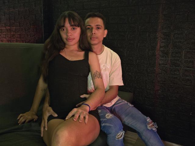 MadissonAndThomas with long brown hair and big tits, wearing a black dress, sits on a couch. Thomas, with short black hair and a tattoo on his arm, hugs her.