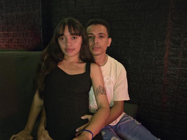 MadissonAndThomas with long brown hair, black dress, and small tits, sits next to Thomas with short brown hair, white tee, and visible tattoo, in a dimly lit room.