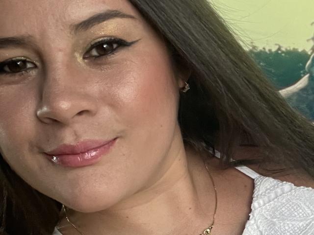 LoliFlirtAti has shiny brown eyes, black winged eyeliner, gold eyeshadow, pink glossy lips with a lip ring, and a white textured top, looking at the camera.