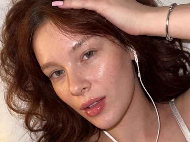 CleoErgaX has shiny, slightly freckled skin, brown wavy hair, pink glossy lips, white earbuds, and a silver bracelet on her right hand, with her hand in her hair.