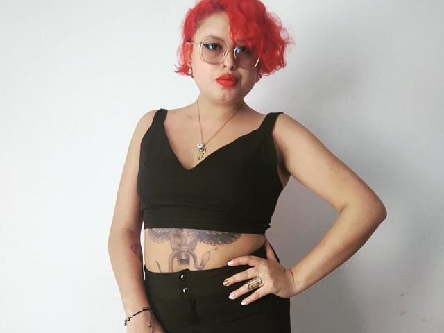 RubiBabee with bright red curly hair, glasses, black crop top, showing a tattoo of a bird below her tits, black high-waisted pants, hand on hip, white background.