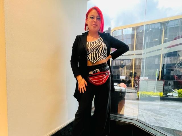 RubiBabee has bright red hair, black pants, a zebra print top, black jacket, and a red chain purse, standing by a glass window, looking confident.