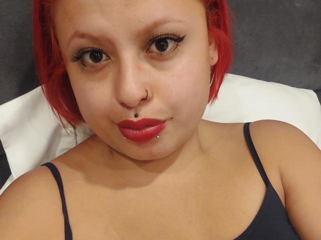 Close-up of RubiBabee with red hair, light brown skin, and dark eyeliner. She has a nose ring and a lip piercing on her bottom lip. She wears a dark spaghetti-strap top. Her lips are painted bright red. The image focuses on her face, showing a slight, confident smile. The background is blurred, emphasizing her facial features and piercings. The composition is centered, highlighting her upper torso and face.
