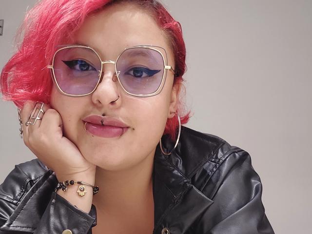 RubiBabee has pink-tipped hair, gold-rimmed glasses, black leather jacket, nose ring, lip ring, tongue piercing, hoop earrings, and black eyeliner, with hand on cheek.