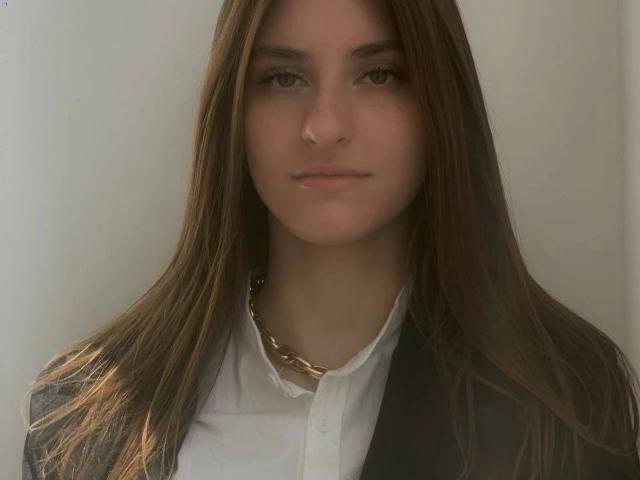 GeorgiannaWow with long brown hair, wearing a black blazer over a white shirt, and a gold chain necklace, has a neutral expression, looking straight at the camera.