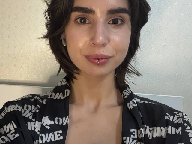 TameikaMacclellan with short dark hair, brown eyes, slight smile, black patterned shirt, small nose piercing, and subtle makeup, looking at camera with a calm expression.