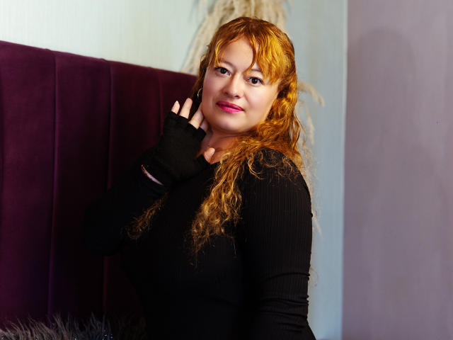 SusanNovak with curly red hair, black ribbed long-sleeve shirt, fingerless black glove, pink lipstick, smiling, hand near face, purple velvet headboard in background.