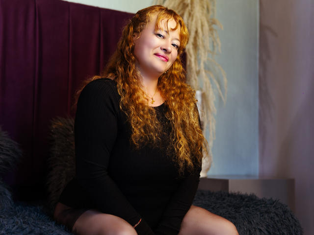 SusanNovak with curly red hair, black long-sleeve shirt, and pink lipstick, sitting on a bed with dark purple headboard, smiling, showing her tits, and looking cute.