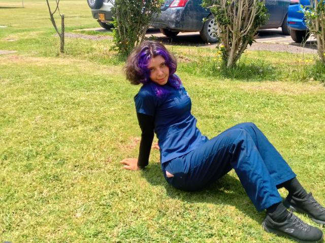 DarckVelvet sits on grass with her back partially on the ground. She has purple-dyed hair and wears a blue medical uniform with black sleeves. She leans on her right arm, legs bent at the knees, and looks at the camera with a slight smile. She's wearing black boots. The grass is green, and there's a small tree and a blue car in the background. Her hair contrasts with the blue uniform, adding a pop of color. She's relaxed, with a casual, almost playful posture.