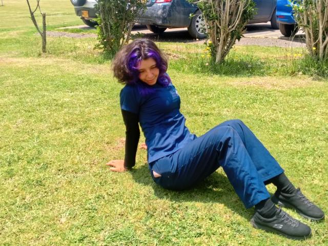 DarckVelvet with purple hair, sitting on grass in blue scrubs and black shoes, black long-sleeve shirt, looking at camera, legs bent, hands on ground, sunny day, casual.
