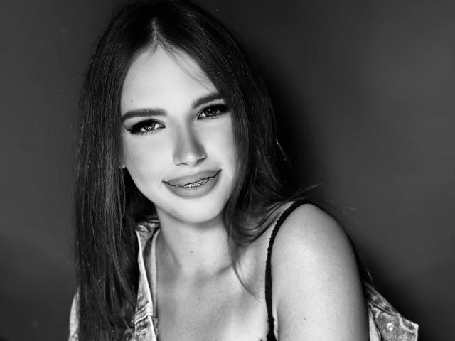 JuanaGoldmann has long dark hair, black eyeliner, and a slight smile with visible metal teeth. She's wearing a denim jacket with black straps. Black and white photo.