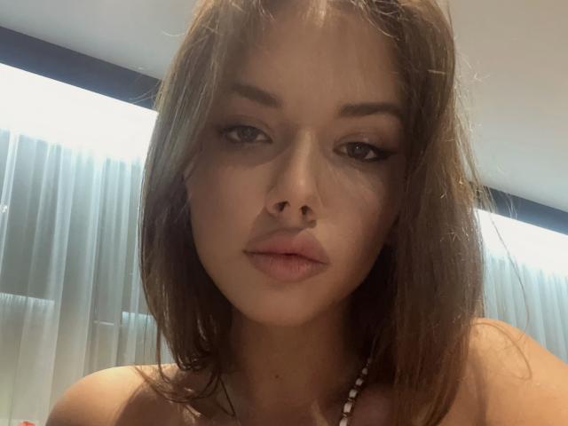 JuanaGoldmann has brown, shoulder-length hair, full lips, and slightly parted pink lips. She's wearing a necklace, and her face is centered in the image with a neutral expression.