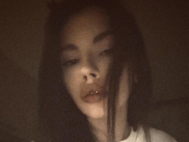 Blurry photo of JuanaGoldmann with dark, wavy hair, slightly parted lips, and a soft, shadowy face, giving a sultry, half-lidded expression. Her skin is light, and she's wearing a white shirt.