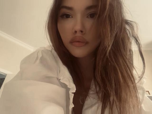 JuanaGoldmann with long brown hair, wearing a white shirt, looking down at the camera with a slight pout, her tits barely visible, in a softly lit room.