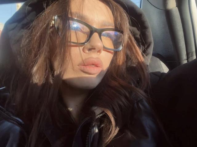 JuanaGoldmann with puffy lips, wearing black glasses and a black hoodie, sunlight in her brown hair, sitting in a car, looking slightly to the side.