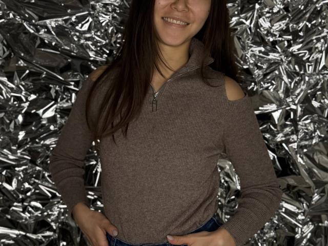 DeniseStringari with brown hair, smiling, wearing a ribbed, zipper-front, brown top with cut-out shoulders, hands on hips, standing in front of silver foil.