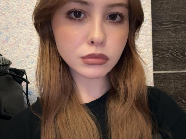 AdinaRosemore has light skin, brown eyes, and long brown hair, wearing a black shirt, with a neutral expression, and subtle pink lipstick. Her nose is small and her lips are full.