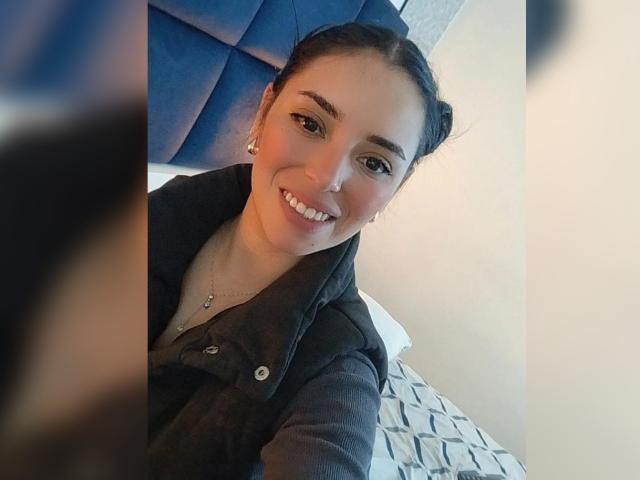 MartinaRey with dark hair in braids, smiling, wearing a black shirt, showing her teeth, earrings, and a necklace, in a bedroom with a blue headboard.