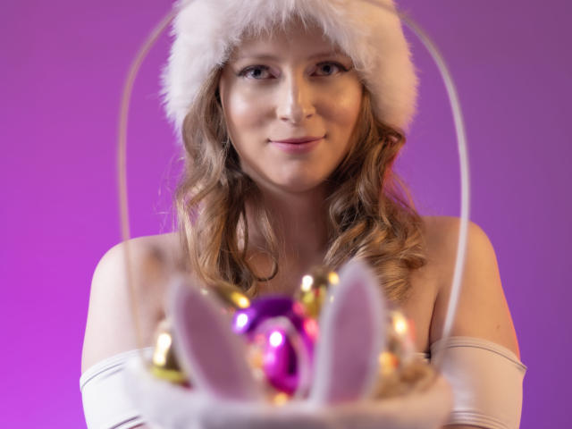 AmbeerWood with wavy brown hair, wearing a white fur hat and white gloves, holding a basket with purple and gold ornaments and white bunny ears, smiling.