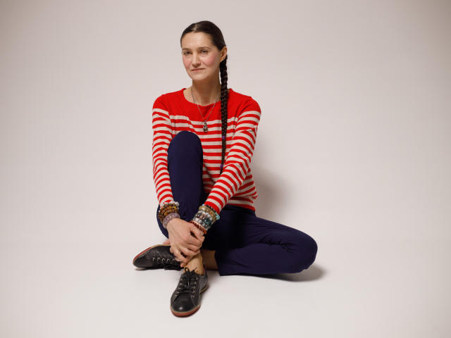 TayaFlavy sits on the floor with her legs crossed, wearing a red and white striped long-sleeve shirt, dark blue pants, and black lace-up shoes. Her long dark hair is braided and she has a subtle smile. She has multiple colorful bracelets on her right wrist. The image is straightforward with a white background, emphasizing her relaxed, casual pose. The stripes on her shirt are bold and evenly spaced.