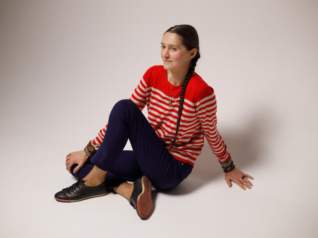 TayaFlavy sits on a plain white floor, legs crossed, right arm resting on the ground, left arm slightly bent. She wears a red and white striped long-sleeve shirt, dark blue pants, and black sneakers. Her brown hair is in a braid, and she has a slight, relaxed smile. Her right wrist has a brown bracelet. The lighting is even, with no visible shadows. She looks slightly to the left.