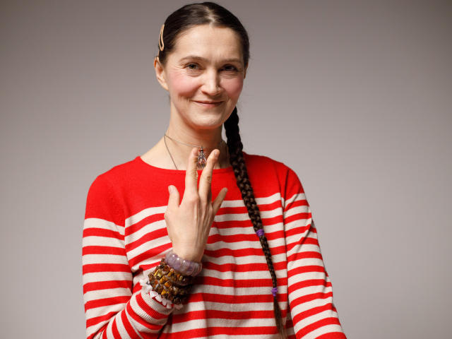 TayaFlavy has a light skin tone and dark brown hair in a single braid. She wears a red and white striped long-sleeve shirt and multiple beaded bracelets on her right wrist. Her right hand is raised, showing a ring with a red stone on her index finger. She has a slight smile and a subtle blush on her cheeks. The image has a neutral gray background, and the composition is centered. The texture of the shirt is slightly loose, and the beaded bracelets add a colorful contrast to the striped pattern.