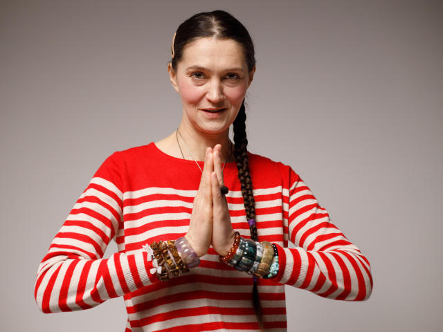 TayaFlavy stands center-frame with hands pressed together in a prayer-like position. She has a fair skin tone, brown hair in a single braid, and wears a red and white striped long-sleeve shirt. Her face shows a slight, calm smile. She has multiple colorful bracelets on both wrists. The image is clear and well-lit, with a neutral gray background. The composition is straightforward, focusing on TayaFlavy's upper body and expression.
