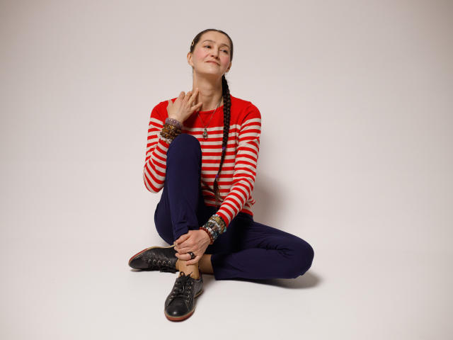 TayaFlavy sitting cross-legged, wearing a red-striped sweater, dark blue pants, black shoes, with braids, touching her neck, looking up with a slight smile.