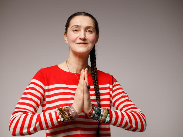 TayaFlavy, a woman with fair skin and brown hair in a braid, stands with hands pressed together in a prayer position. She wears a red and white striped long-sleeve shirt and multiple colorful bracelets on both wrists. Her expression is calm and slightly smiling. The image is a studio portrait with a neutral grey background, highlighting her centered, symmetrical composition. The lighting is even, emphasizing her facial features and the textures of her clothing.