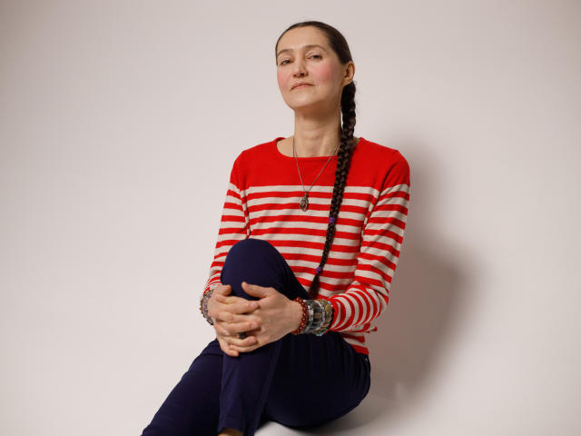 TayaFlavy has brown hair in a braid, wearing a red and white striped shirt, dark pants, and multiple bracelets, sitting with knees up, looking slightly to the side.