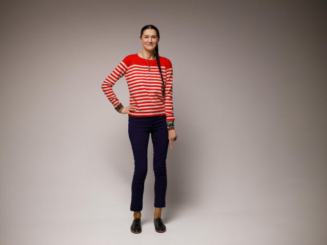 TayaFlavy stands with a slight smile, wearing a red and white striped long-sleeve shirt, dark navy pants, and black loafers. She has a long braid over her right shoulder, and multiple bracelets on her left wrist. The image is well-lit with a neutral grey background. The composition is centered, showing TayaFlavy from head to toe. The textures are smooth, and the colors are vibrant. Her stance is relaxed and confident.