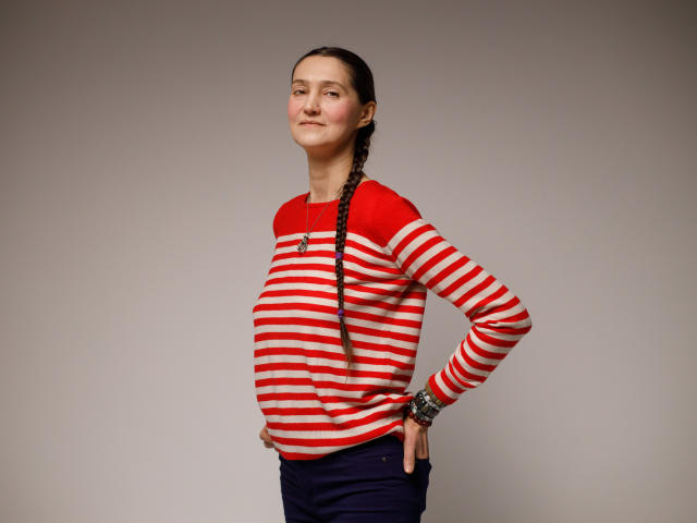 TayaFlavy stands against a plain gray background. She has a fair skin tone, dark braided hair, and a slight smile. She wears a red and white striped long-sleeve shirt and dark blue pants. Her right hand rests on her hip, and she has a black bracelet on her right wrist. The image is well-lit with soft shadows. She has a confident, relaxed posture.