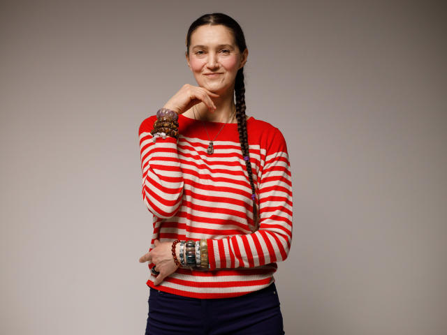 TayaFlavy stands against a plain gray background. She wears a red and white striped long-sleeve shirt with navy pants. Her brown hair is in a single braid falling over her right shoulder. She has a slight smile, with her right hand resting on her chin and her left hand on her stomach. She wears multiple brown bracelets on her right wrist and a ring on her left index finger. The image is straightforward with a soft, even light.