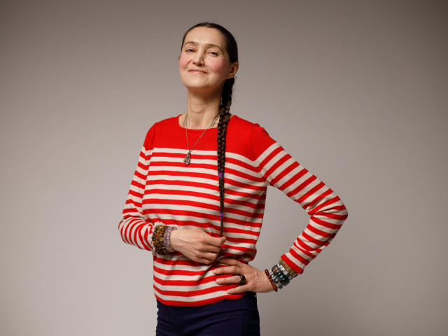 TayaFlavy stands with a slight smile, wearing a red and white striped long-sleeve shirt. Her right hand rests on her hip while her left hand holds a braid of her dark brown hair. She has multiple bracelets on her right wrist, including a colorful beaded one. The shirt has red patches on the shoulders. Her skin is light with a few freckles. The image is well-lit with a soft, neutral background. She exudes a casual, confident vibe.