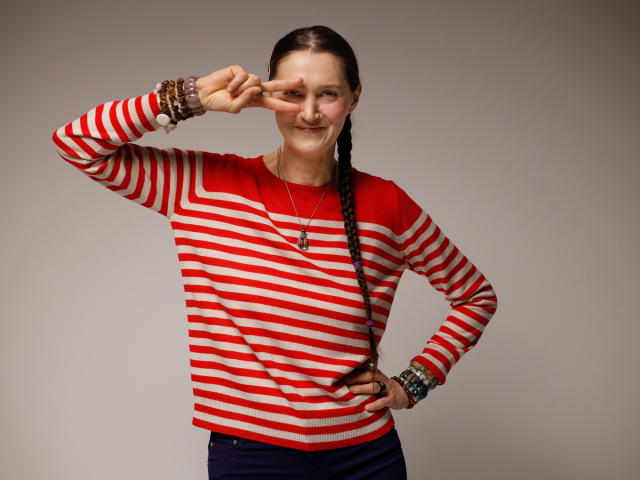 TayaFlavy stands against a plain gray background. She has a fair skin tone and dark brown hair in a single braid. She wears a red and white striped long-sleeve shirt and dark blue pants. Her right hand rests on her hip, and her left hand is raised, making a "peace" sign with her fingers. She has multiple bracelets on her left wrist. She has a slight, confident smile. The lighting is even, highlighting her facial features and clothing.