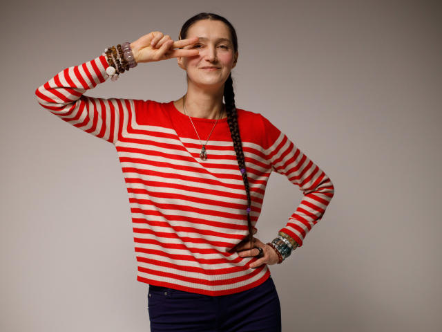 TayaFlavy stands with one hand on her hip and the other making a peace sign near her face. She has a braided pigtails and wears a red and white striped long-sleeve shirt, navy pants, and multiple bracelets on her right wrist. She has a confident, slightly smug expression. The image is a photograph with a simple, neutral background. Her red shirt contrasts with the striped pattern. The lighting is even, highlighting her facial features and the textures of her clothing.