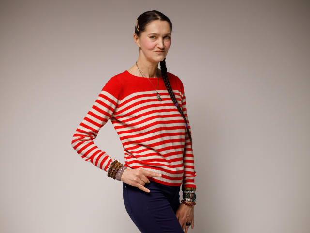 TayaFlavy stands against a plain background, wearing a red and white striped long-sleeve shirt and navy pants. She has a slender build and a long, dark braid that hangs over her right shoulder. Her right hand is on her hip, and she wears multiple colorful bracelets on her left wrist. Her expression is neutral, and she looks slightly to the left. The image is clear and well-lit, with a focus on TayaFlavy's casual, relaxed posture.