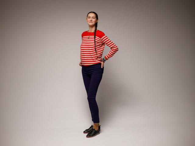 TayaFlavy stands in a neutral, slightly gray studio. She's wearing a red and white striped long-sleeve shirt with a high collar, navy blue skinny pants, and black loafers with red accents. Her dark hair is in a simple braid. She has a relaxed, slightly confident stance with hands on her hips. The image is sharp with a simple, clean composition. Her expression is neutral, and she looks directly at the camera.